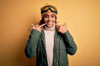 Genç Afro-Amerikan kayakçı kız kar sporu giysisi ve kayak gözlüğü takıyor, gülümsüyor ve parmakları, dişleri ve ağzı ile işaret ediyor. Diş sağlığı konsepti.