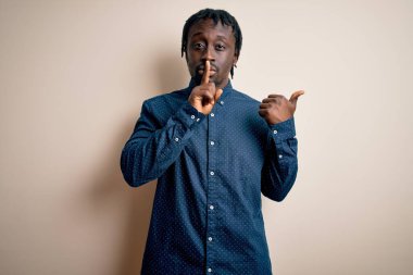 Beyaz arka planda dikilmiş, elleri yandan işaret eden parmaklarıyla sessiz olmak isteyen, gündelik gömlekler giyen genç, Afro-Amerikan bir adam. Sessizlik ve gizli konsept.