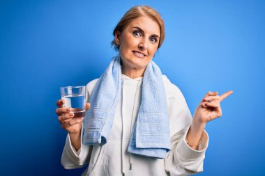 Orta yaşlı güzel sarışın sporcu kadın ferahlatıcı bir bardak su içerek elini ve parmağını kenarda tutarak mutlu bir şekilde işaret ediyor.