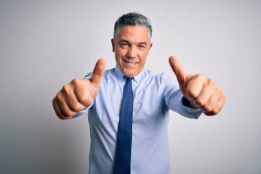 Orta yaşlı, yakışıklı, gri saçlı, şık gömlekli ve kravatlı bir iş adamı. Elle olumlu jest yapmayı onaylıyor. Başparmağıyla gülümsüyor ve başarı için seviniyor. Kazanan jest.