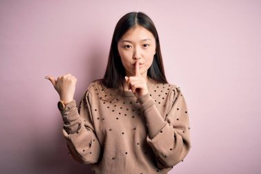 Pembe arka planda zarif bir süveter giyen genç, güzel Asyalı bir kadın parmaklarını yana doğru uzatarak sessiz olmak istiyor. Sessizlik ve gizli konsept.