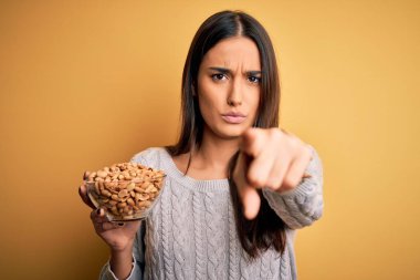 Elinde fındık kabı olan genç ve güzel esmer kadın izole edilmiş sarı arka planda kamerayı işaret ediyor ve sen, el işareti, ön taraftan pozitif ve kendinden emin bir hareket.