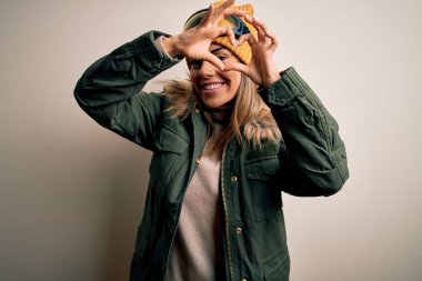 Genç esmer kayakçı kadın kar giysileri ve kayak gözlükleriyle beyaz arka planda kalp şeklinde elleri ve parmaklarıyla tabelanın içinden bakıp gülümsüyor.