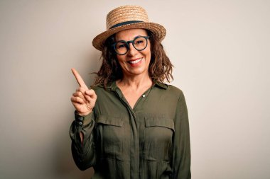 Orta yaşlı esmer kadın gözlüklü ve şapkalı. İzole edilmiş beyaz arka planda duruyor. Yüzünde kocaman bir gülümseme var..