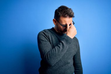 Mavi arka planda duran sıradan süveter ve gözlük takan genç yakışıklı adam yorgun burnu ve gözleri ovuşturarak yorgunluk ve baş ağrısı hissediyor. Stres ve hüsran kavramı.