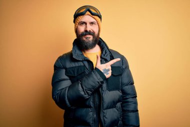 Sakallı, kel yakışıklı kayakçı kar maskesi takıyor ve kayak gözlükleri takıyor. Yüzünde mutlu ve doğal bir ifade olan yüzü, eli ve parmağı yana dönük gülümsüyor.