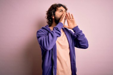 Genç, yakışıklı, sakallı, pembe arka planda sıradan bir eşofman giyen, elleri ağzında sinirli bir şekilde bağıran yakışıklı bir adam.