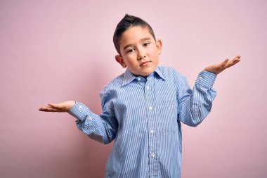 Pembe izole edilmiş arka plan üzerinde duran zarif tişörtlü küçük bir çocuk. Kolları ve elleri havada, kafası karışmış bir yüz ifadesi. Şüphe kavramı.