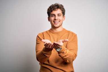 Sarışın, kıvırcık saçlı, beyaz arka planda sıradan bir süveter giyen elleri avuçlarında gülümseyen ya da el hareketi yapan genç bir adam. Tutun ve koruyun