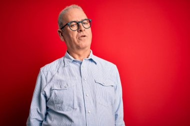 Orta yaşlı yakışıklı, beyaz gömlekli ve gözlüklü bir adam kırmızı arka planda uykulu ve yorgun görünüyor, yorgunluktan yorgun ve akşamdan kalma, sabahları tembel gözler..