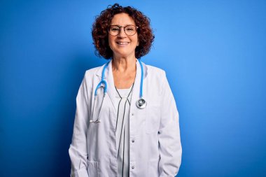 Orta yaşlı, kıvırcık saçlı, palto giyen ve mavi arka planda steteskop takan mutlu ve havalı bir gülümsemesi olan bir kadın. Şanslı kişi..