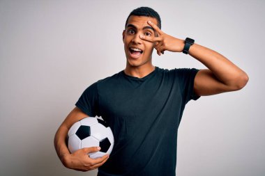 Yakışıklı Afro-Amerikan adam futbol topunu beyaz arka planda tutuyor barış sembolü yapıyor parmakları yüzünde, neşeyle gülümsüyor zaferi gösteriyor.