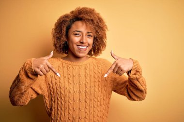 Young beautiful African American afro woman with curly hair wearing casual sweater looking confident with smile on face, pointing oneself with fingers proud and happy.