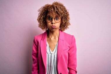 Genç Afro-Amerikalı iş kadını, pembe arka planda duran gözlüklü komik yüzlü, pofuduk yanaklı. Havayla şişmiş ağız, çılgın bir ifade..