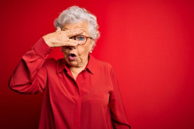 Son sınıf güzeli, gri saçlı, günlük gömlek ve gözlük takan, kırmızı arka planda, şokla yüzünü ve gözlerini kapatan, utangaç ifadesiyle parmaklarının arasından bakan bir kadın..