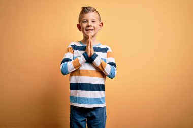 Mavi gözlü, sarı arkaplan üzerine çizgili tişört giyen, elleri bağlı, af dileyen, kendinden emin, gülümseyen küçük beyaz bir çocuk..