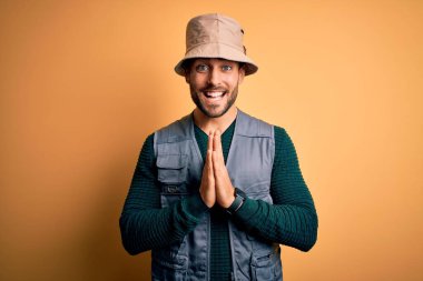 Tatilde sakallı yakışıklı bir turist sarı arka planda kaşif şapkası takıyor elleriyle birlikte dua ediyor, af diliyor kendinden emin bir şekilde gülümsüyor..