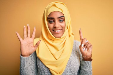 Genç ve güzel Afro-Amerikalı kız izole edilmiş sarı arka plan üzerine Müslüman başörtüsü takıyor ve altı numarayı gösterirken kendinden emin ve mutlu bir şekilde gülümsüyor..