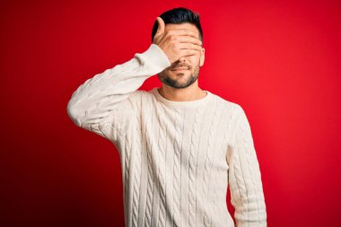 İzole edilmiş kırmızı arka plan üzerinde duran, sıradan beyaz süveter giyen genç yakışıklı bir adam. Gülümsüyor ve elleri yüzünde şaşkınlıktan gözleri kapatıyordu. Kör kavram.