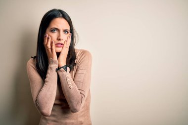 Mavi gözlü genç esmer kadın izole edilmiş beyaz arka plan üzerine günlük süveter giyiyor. Yüzü, depresyonu ve üzüntüyü örten eller, sorun yüzünden üzgün ve tedirgin.