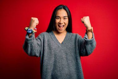 Genç ve güzel Asyalı kadın, gündelik süveter giyip izole edilmiş kırmızı arka planda dikiliyor. Şaşırmış ve şaşırmış bir şekilde kollarını kaldırıp, gözlerini açarak başarıyı kutluyor. Kazanan konsept.