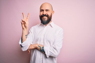 Soyutlanmış pembe arka plan üzerinde zarif bir tişört giyen sakallı yakışıklı kel bir adam. Kameraya gülümsüyor ve zafer işareti yapıyor. İki numara..