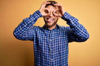 Sarışın, sakallı ve mavi gözlü genç bir işadamı sarı arka planda gömlek giyiyor. Dürbün gibi hareket ediyor, gözleri parmaklarından bakıyor. Çılgın ifade.