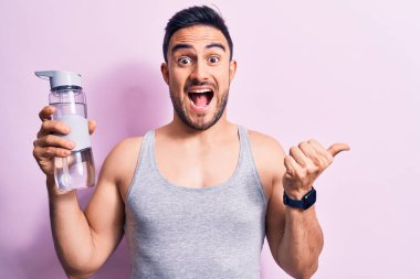 Sakallı, kolsuz tişörtlü, su şişesi giyen genç yakışıklı sporcu başparmağıyla yan tarafa doğru gülümsüyor ve ağzı açık gülümsüyor.