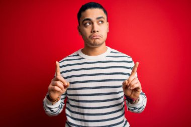İzole edilmiş kırmızı arka planda dikilen sıradan çizgili tişört giyen genç Brezilyalı adam üzgün ve üzgün görünüyor, parmaklarıyla yönü gösteriyor, mutsuz ve depresif..