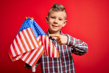 Küçük Amerikalı vatansever beyaz çocuk kırmızı arka planda ABD bayrağını tutuyor. Elleri ve parmaklarıyla işaret ederek çok mutlu.