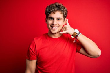 Sarışın, kıvırcık saçlı, kırmızı arka planda sıradan tişörtler giyen, el ve parmaklarıyla telefonla konuşan genç, yakışıklı bir adam. İletişim kavramları.