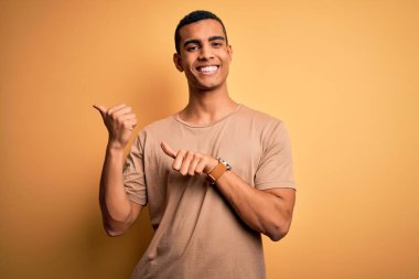 Genç, Afro-Amerikalı, günlük tişört giyen, sarı arka planda duran, el ve baş parmaklarıyla arkasını gösteren, kendinden emin gülümseyen bir adam.