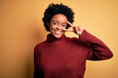 Kıvırcık saçlı, balıkçı yaka kazak giyen, yüzünde parmaklarıyla barış sembolü çizen, gülümseyen, zafer gösteren güzel Afro-Amerikan kadın.