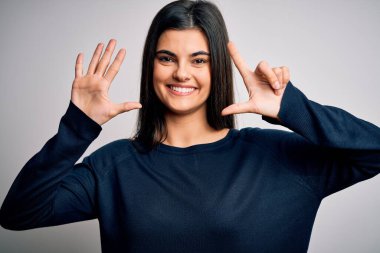 Genç, güzel, esmer, sıradan bir süveter giyen, beyaz arka plan üzerinde duran, yedi numaralı parmaklarıyla işaret eden, kendinden emin ve mutlu bir şekilde gülümseyen bir kadın..