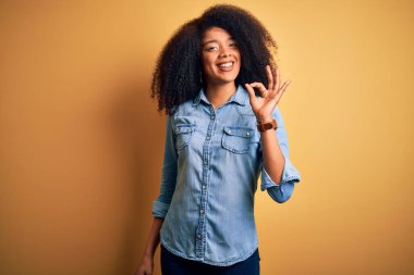 Afro saçlı, sarı izole edilmiş arka planda duran güzel Afro-Amerikalı kadın gülümsüyor ve el ve parmaklarıyla 