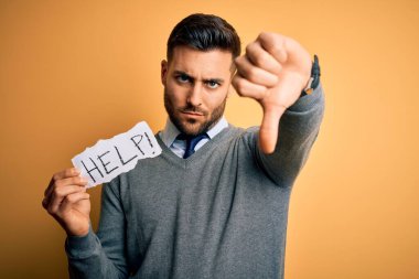 Aşırı çalışan genç iş adamı sarı arka planda, kızgın yüzlü, olumsuz işaretli, baş parmakları aşağıda, reddedilme kavramından hoşlanmadığını gösteren bir kağıt tutarak yardım istiyor.