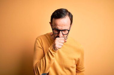 İzole edilmiş sarı arka plan üzerinde günlük süveter ve gözlük takan orta yaşlı beyaz bir adam kendini iyi hissetmiyor ve soğuk algınlığı veya bronşitin belirtisi olarak öksürüyor. Sağlık hizmeti kavramı.