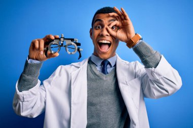 Yakışıklı afro-amerikan optik adam mavi arka planda optometri gözlüğü tutuyor gülümseyen yüzüyle tamam işaret ediyor parmaklarının arasından bakıyor
