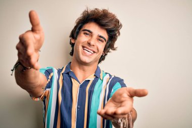 Yaz tatilindeki genç adam, renkli tişörtler giyip izole edilmiş beyaz arka planda dikiliyor ve kameraya sarılıp gülümsüyor. Mutluluğu kucaklayan neşeli bir ifade..