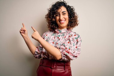 Genç ve güzel kıvırcık Arap kadın, çiçekli tişört giyiyor, izole edilmiş beyaz arka planda duruyor, gülümsüyor ve kameraya bakıyor, iki eliyle ve parmaklarıyla yan tarafa bakıyor..