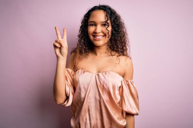 Kıvırcık saçlı, rahat tişörtlü pembe arka planda duran mutlu yüzüyle kameraya gülümseyen parmaklarıyla zafer işareti yapan genç bir kadın. İki numara..