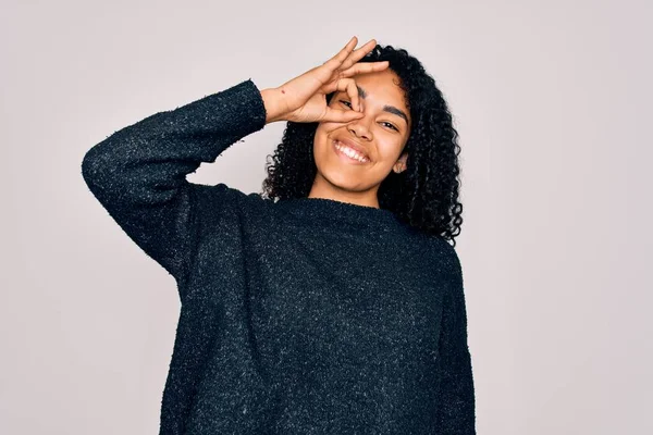 Young african american curly woman wearing casual sweater standing over white background doing ok gesture with hand smiling, eye looking through fingers with happy face.