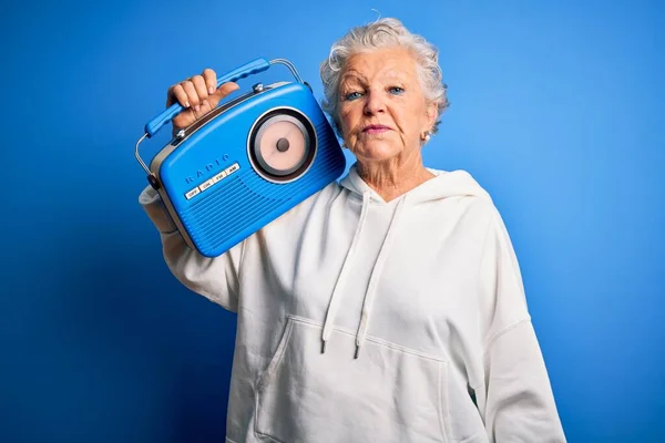 年长的漂亮女人拿着老式收音机 站在孤零零的蓝色背景上 脸上带着自信的表情 严肃地思考着 — 图库照片