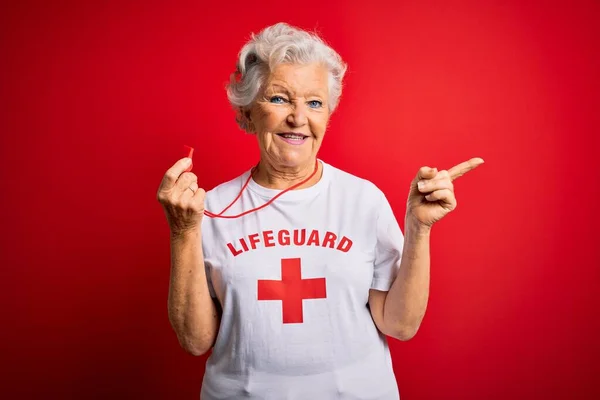 赤い十字のTシャツを身に着けているシニア美しい白髪のライフガードの女性は 幸せと自然な表情で手と指を横に向けて笑顔で明るい笛を使用しています — ストック写真