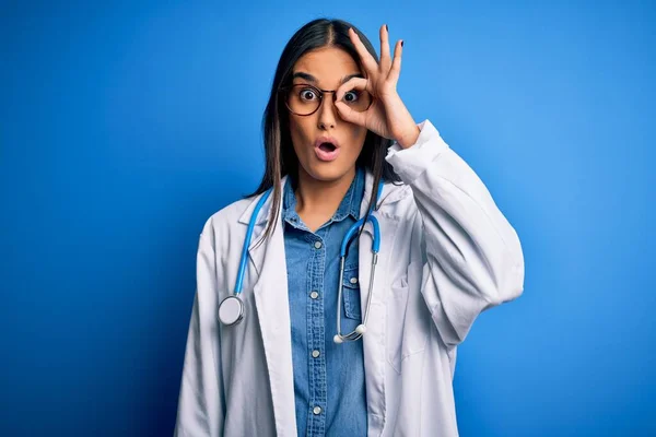 青い背景の上に聴診器と眼鏡をかけている若い美しい医師の女性は 驚きの顔でショックを受けたOkジェスチャーを行うと 目を指で見て 不信仰の表現 — ストック写真