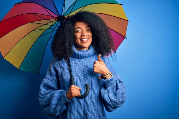 Young african american woman with afro hair under colorful umbrella for winter weather rain happy with big smile doing ok sign, thumb up with fingers, excellent sign