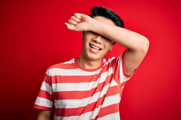 Young handsome chinese man wearing casual striped t-shirt standing over red background covering eyes with arm smiling cheerful and funny. Blind concept.