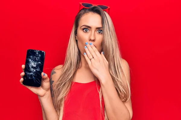 Young beautiful blonde woman holding broken smartphone showing cracked screen covering mouth with hand, shocked and afraid for mistake. Surprised expression