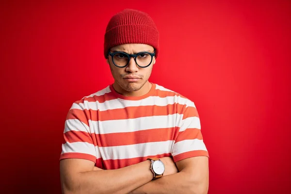 孤立した赤の背景にカジュアルなストライプのTシャツと眼鏡を身に着けている若いブラジル人男性は懐疑的で緊張し 両腕を組んで顔に不承認の表現 負の人 — ストック写真