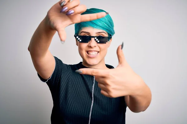 Young woman with blue fashion hair wearing thug life sunglasses over white background smiling making frame with hands and fingers with happy face. Creativity and photography concept.
